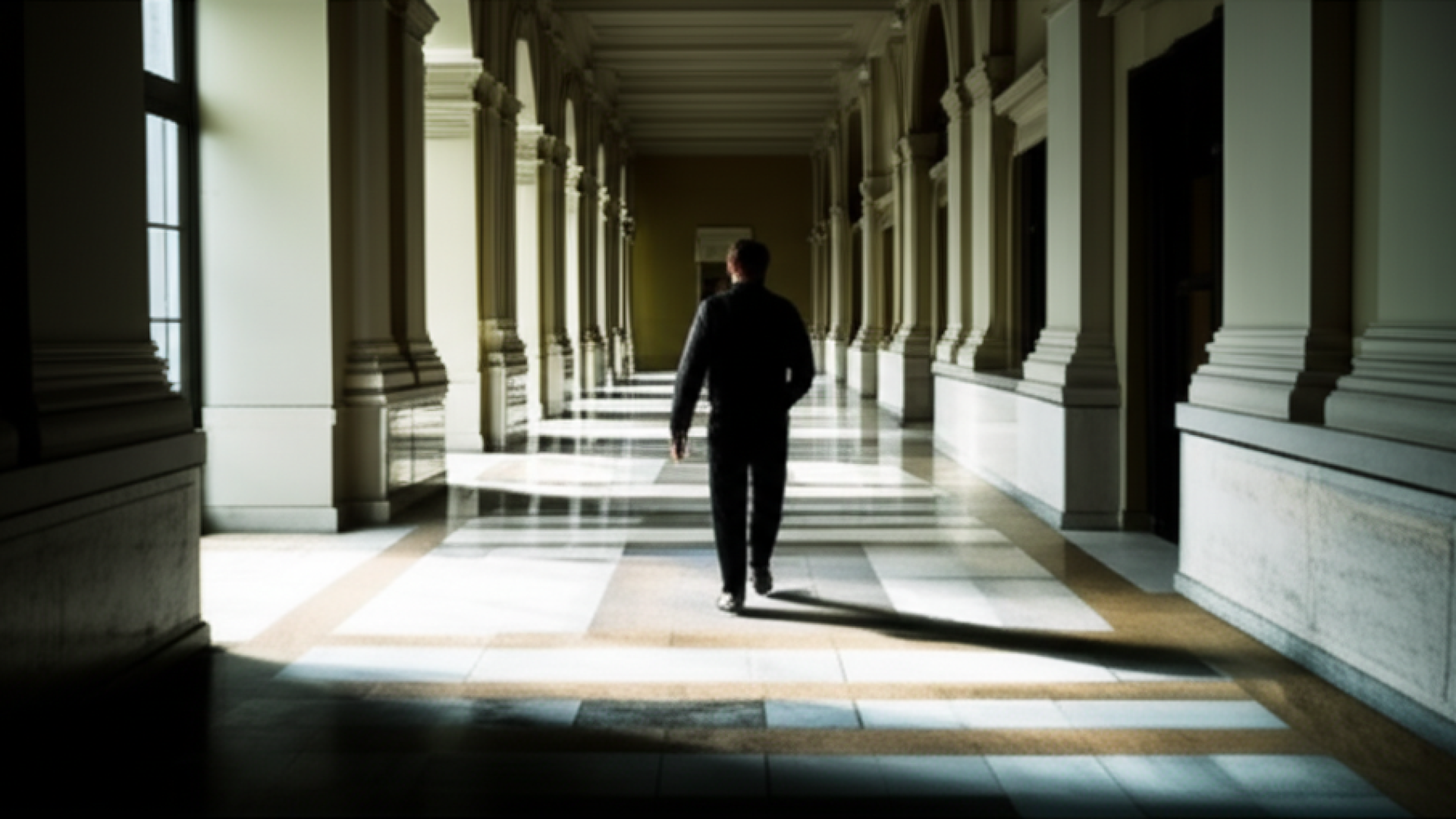 A wide shot of a solitary man walking away from the camera down a grand, empty hallway with marble f